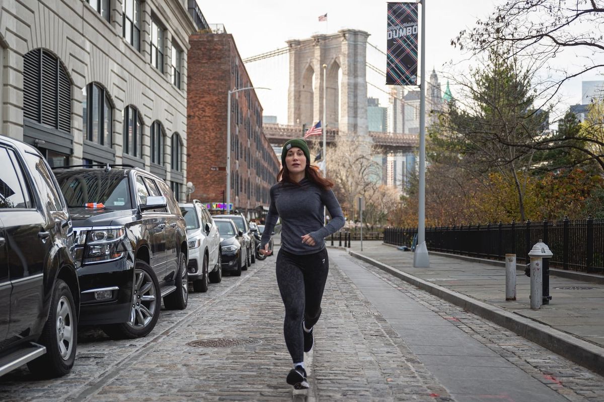 A woman running down a street in a downtown city with a bridge in the background and cars lining the street