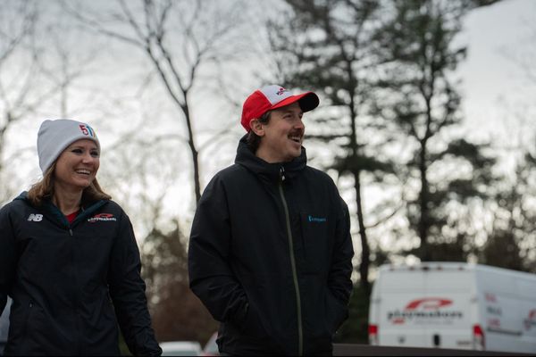 A woman and a man dressed in warm jackets and hats standing outside in front of a white van and trees without their leaves
