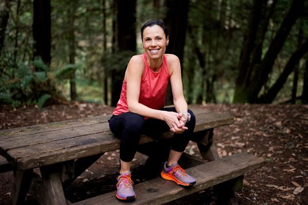 A woman wearing black tights, a peach tank top and trail running shoes sitting on top of a picnic table with forest trees in the background