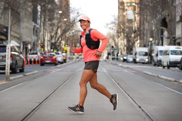 A woman in running shorts, a pink long-sleeve shirt, and a pink hat running across a downtown city street and smiling for the camera