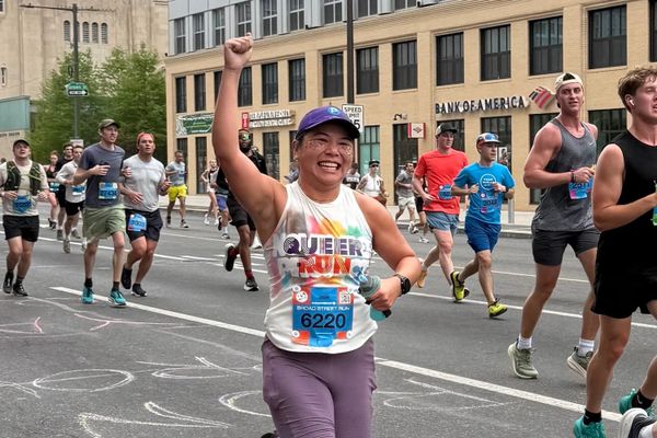 A woman runner in a white tank top, purple shorts, and hat, smiling and raising her fist in excitement while running in a race along a downtown street