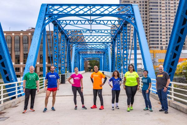 Eight people in brightly colored clothes, posing outside in a city by standing on a walkway with a blue bridge rails above them