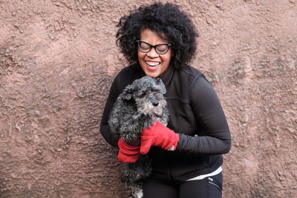 A woman laughing, wearing a black running jacket and red mittens, and holding a small dog