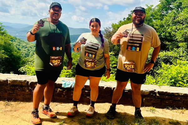 Two men and a woman standing outside wearing running apparel and posing with race medals, with tree-covered hills in the background