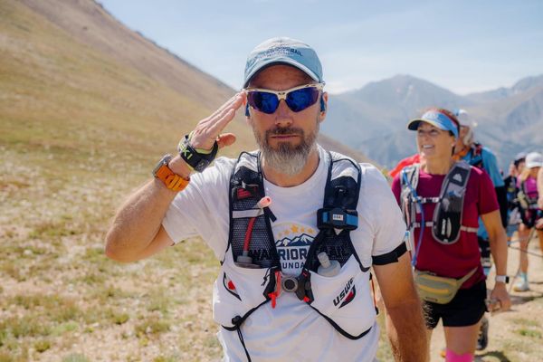 A man giving a salute, wearing a hat, sunglasses, and a running vest while on a trail in the mountains with other runners behind him