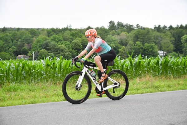 A woman riding a triathlon bike on a road with green trees and a corn field in the background