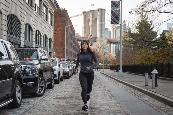 A woman running down a street in a downtown city with a bridge in the background and cars lining the street