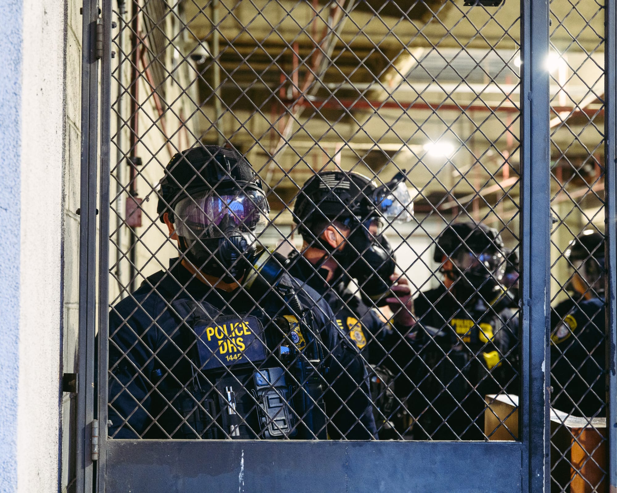 Four police officers stand behind a metal gated door. They are allo wearing space-age protective gear. 