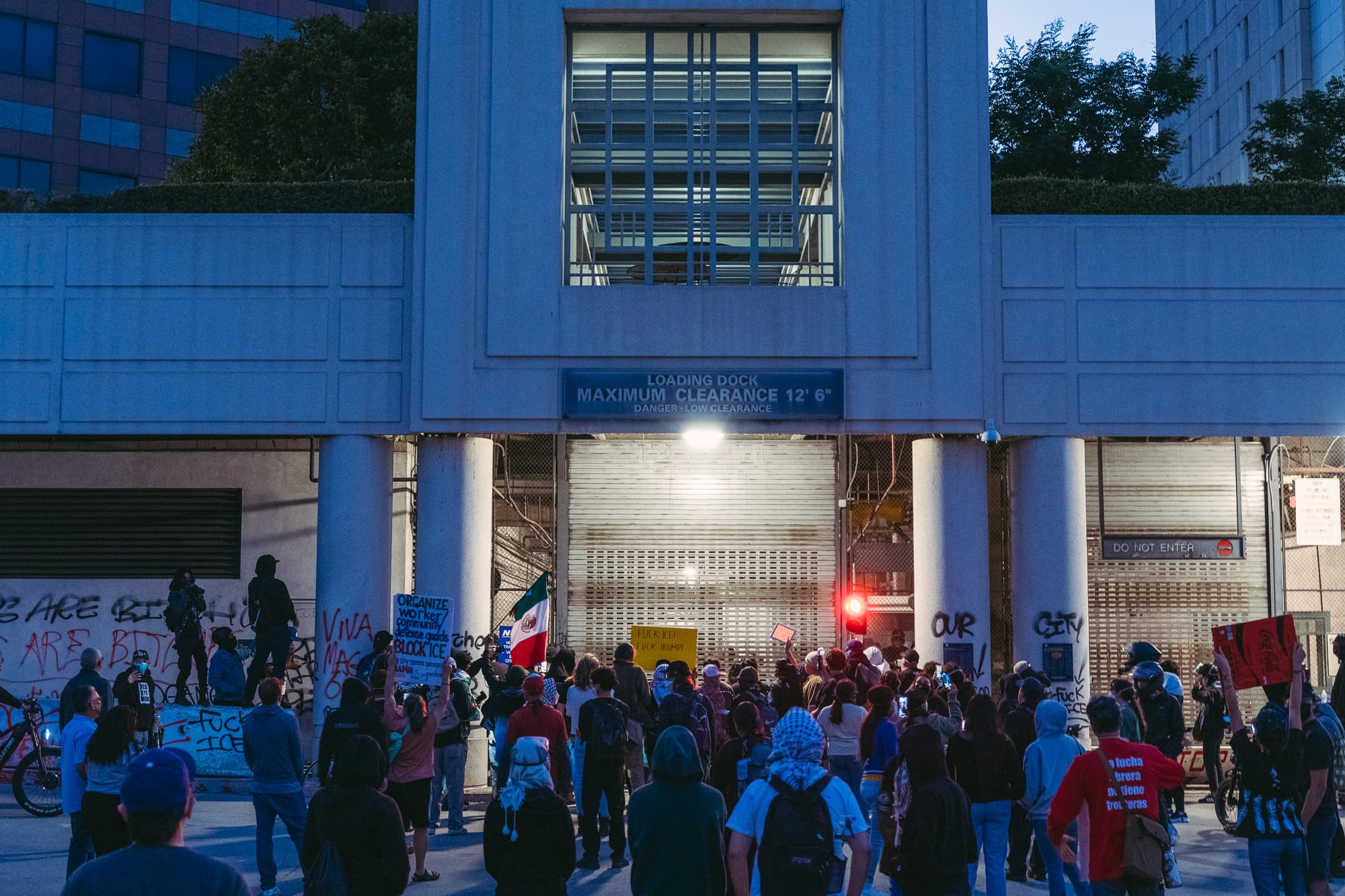 A building at dusk. A garage door light is on. A group of people are spread out on the driveway. 