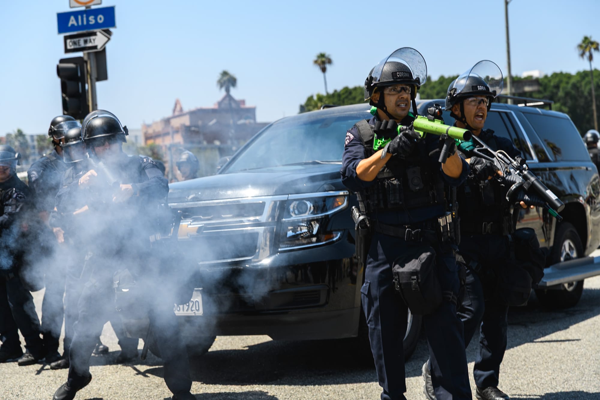 Three cops stand infront of a SUV. All three of them in riot gear and guns. Smoke is coming out of one them after an officers fires it. 