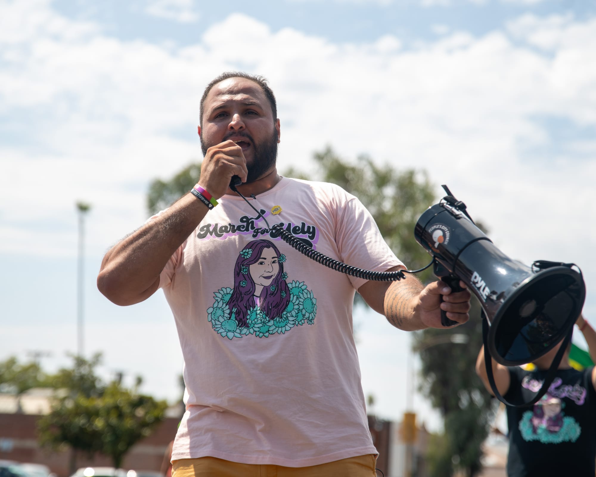 A man uses a megaphone wearing a pink shirt that says March for Mely. 