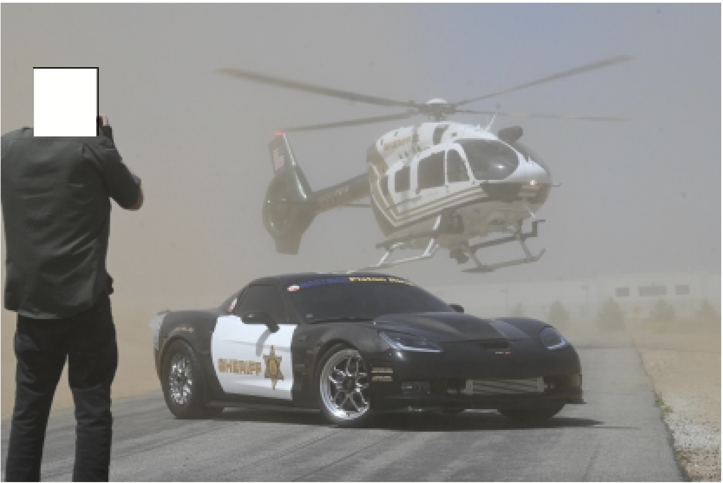 A helicopter hovers in the background of a parked Corvette that looks like a police car. A person with a white box over his head takes a photo.
