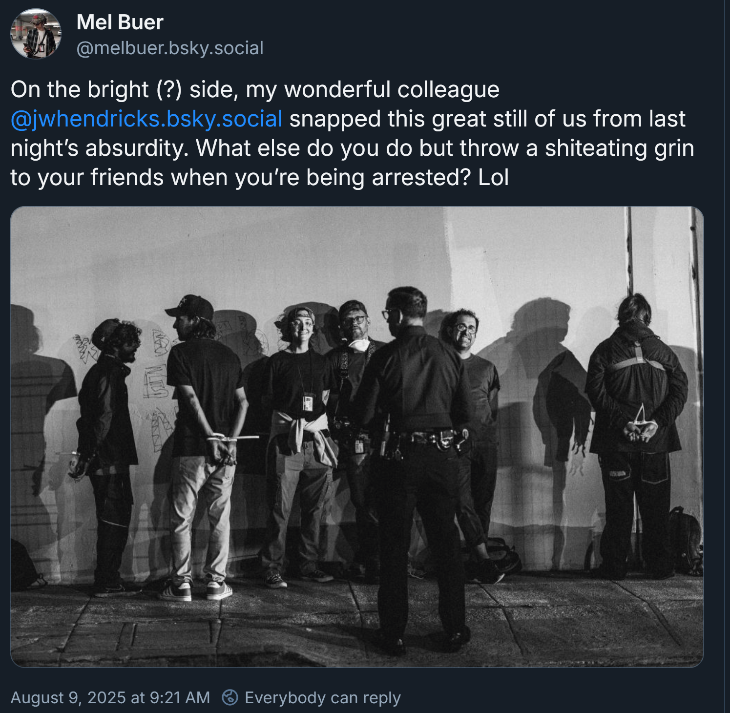 A social media post with a photo of people lined up against the wall, including three people facing and smiling the camera. The post reads On the bright (?) side, my wonderful colleague JW Hendricks snapped this great still of us from last night's absurdity. What else do you do but throw a shiteating grin to your friends when you're being arrested? lol