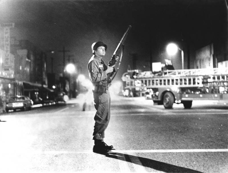 A solider stands with a rifle in his hand. Behind him is a fire truck.