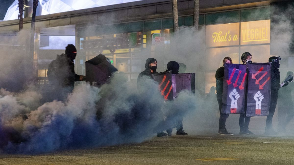 People holding shields and umbrellas are covered in black smoke. The signs have red arrows pointing down and a raised fist.