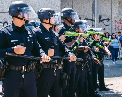 A row of police officers wearing helmets with visors stand next to each other. Several are holding batons, others are holding guns painted green, and they're pointing at people.