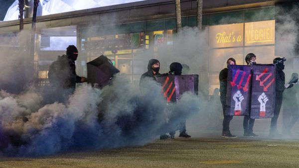 People holding shields and umbrellas are covered in black smoke. The signs have red arrows pointing down and a raised fist.