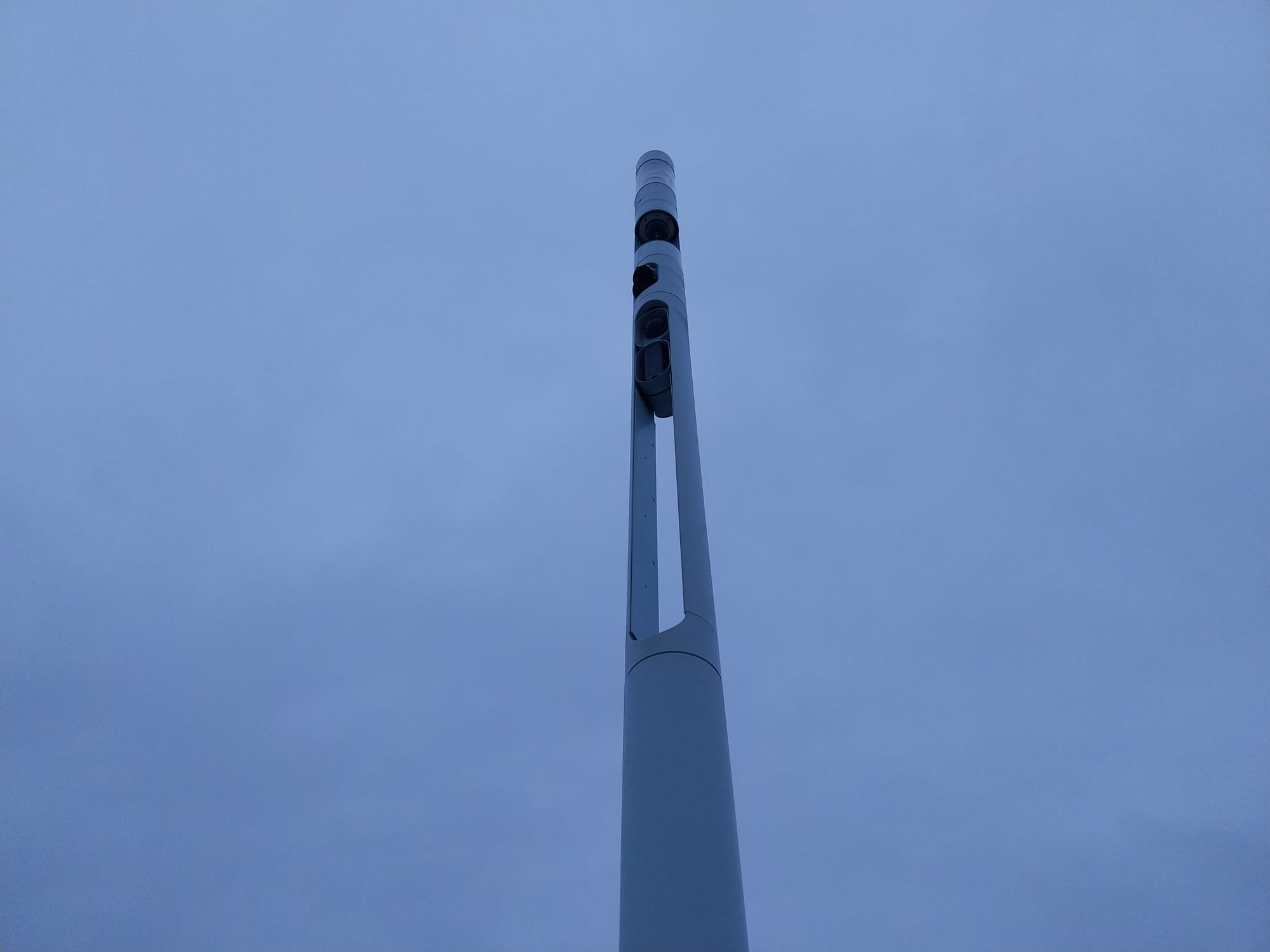 Photo of a grey-blue light post against a grey-blue sky. The light post is thin and cylindrical, and has a number of dark-coloured modules along the length of its body, including a camera.