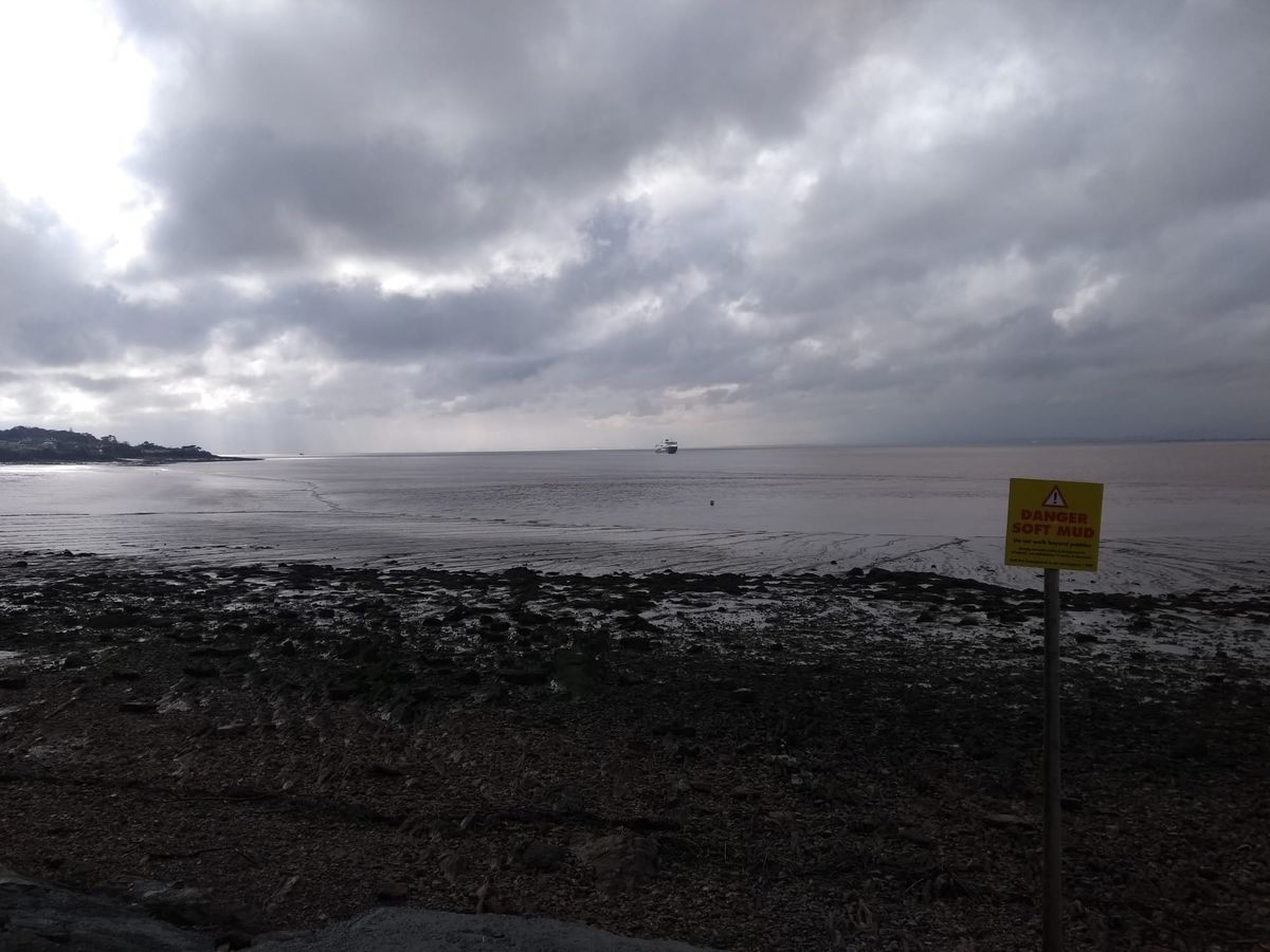 Photo of a beach and a bright but cloudy sky. There is a ship far away on the horizon, and a yellow sign in the foreground that says "Danger Soft Mud."