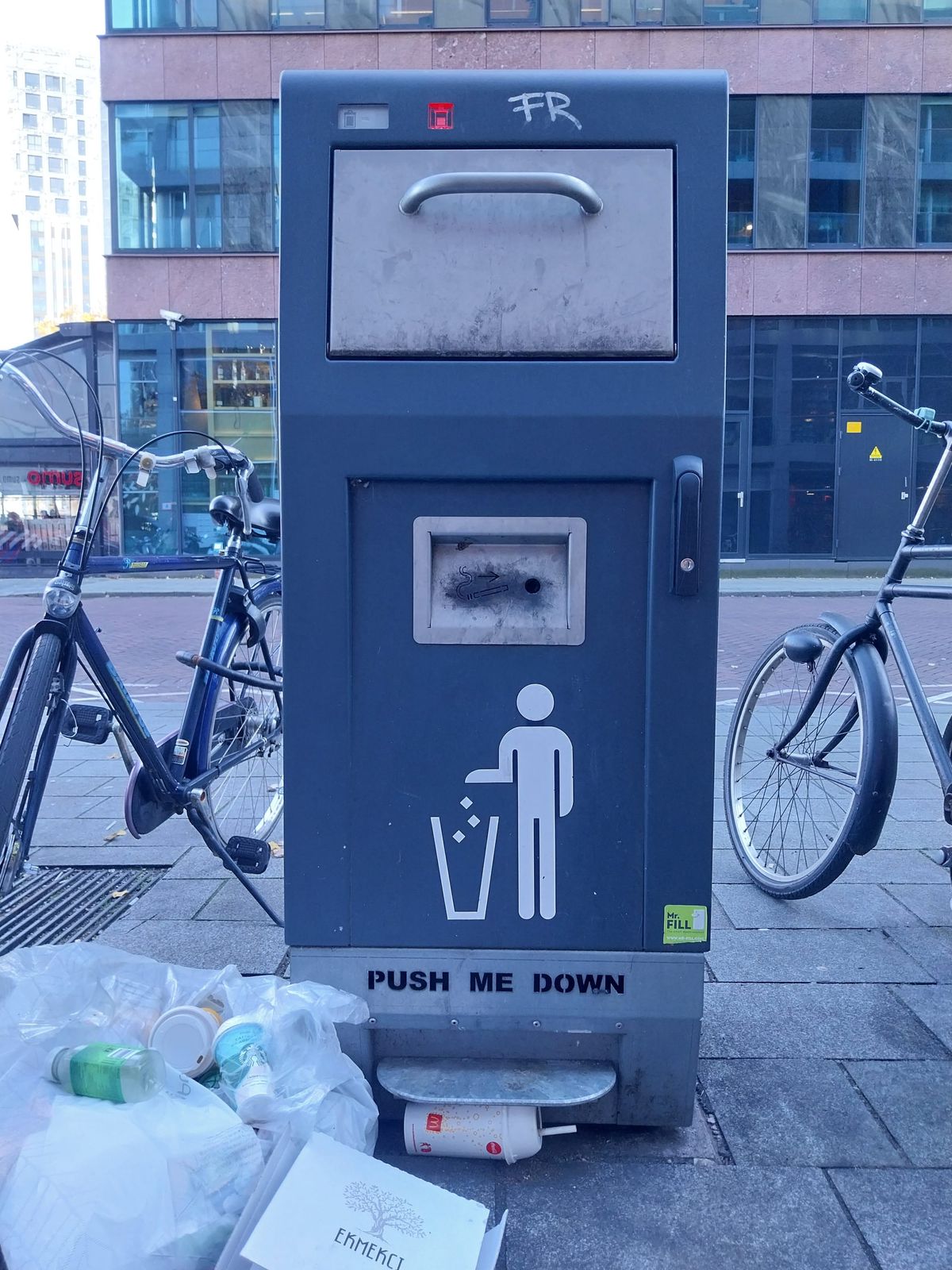 A photo of a smart garbage can on a street, surrounded by garbage. The red indicator light on the garbage can indicates that it is locked.
