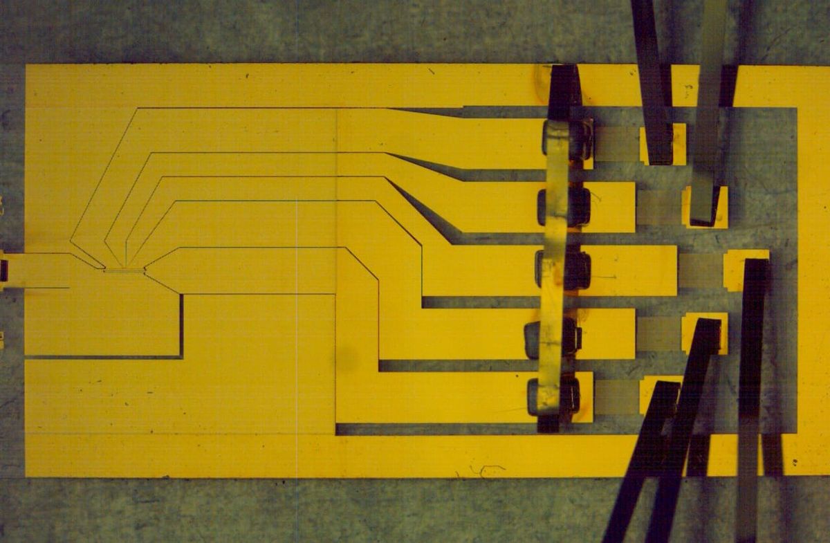 A slightly low-contrast image of a sheet of yellow material with paths cut out of it. At the ends ofthe paths, there are strips of sheet metal sticking up and out.