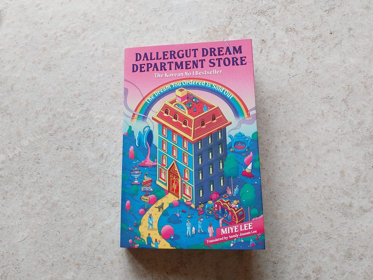 A paperback book on a concrete background. The book is brightly-coloured and depicts a whimsical building. The book is DallerGut Dream Department Store by Miye Lee.