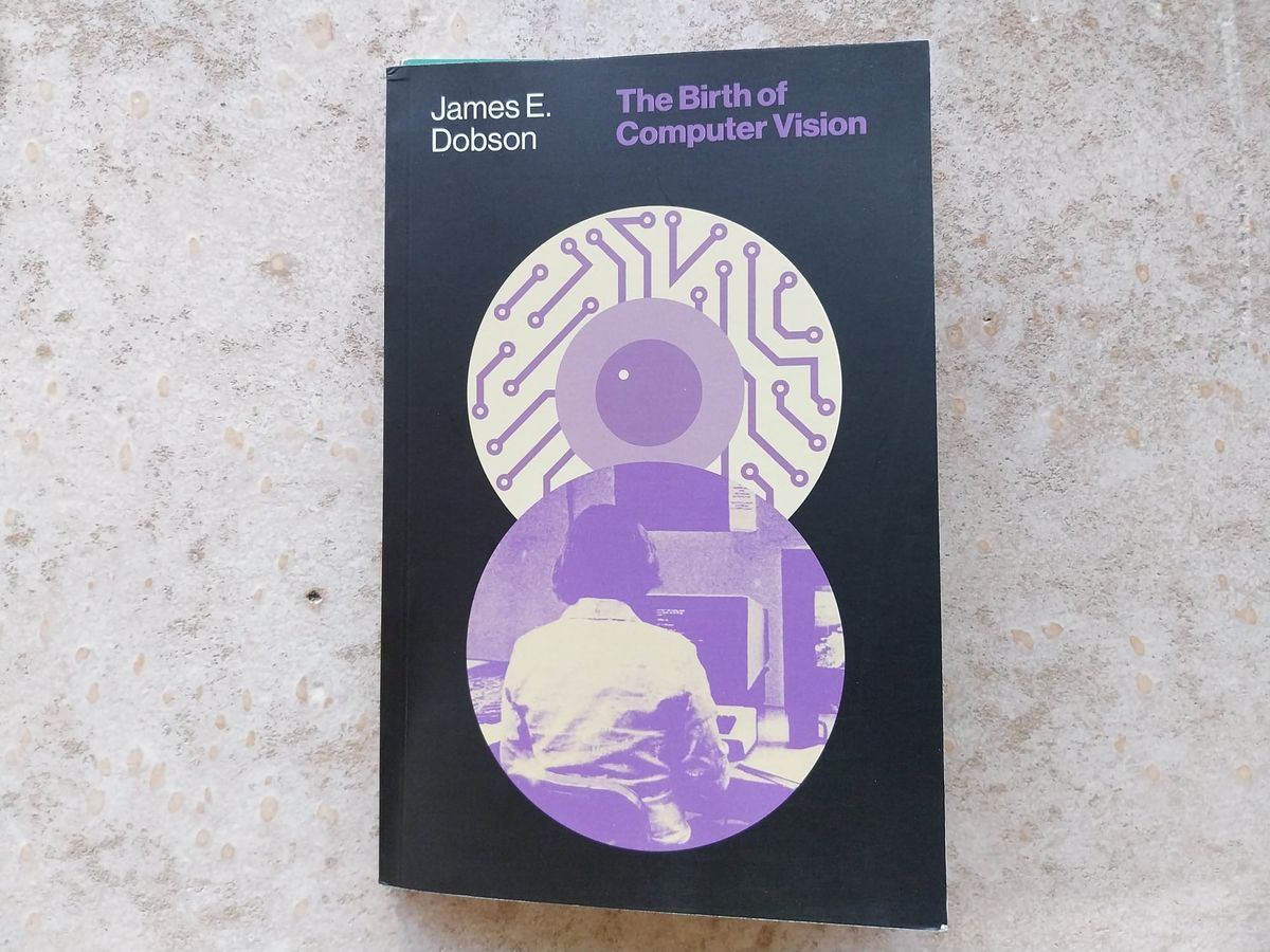On a concrete floor, a book. The book is The Birth of Computer Vision by James E. Dobson. It has a black cover with some purple-tinted, slightly abstract images representing computation.