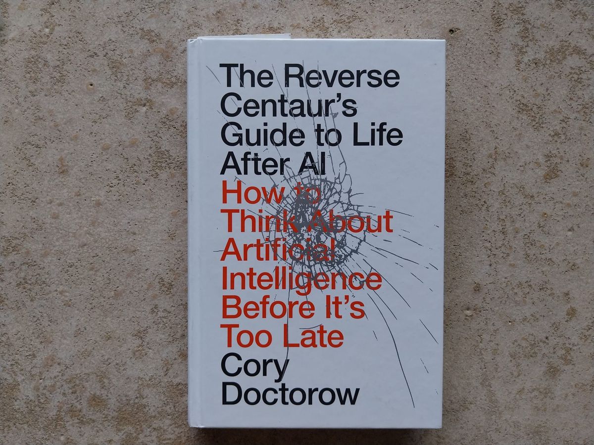 Photo of a book on a concrete floor. The book is white, with red and black text on the cover. It is "The Reverse Centaur's Guide to Life After AI" by Cory Doctorow.