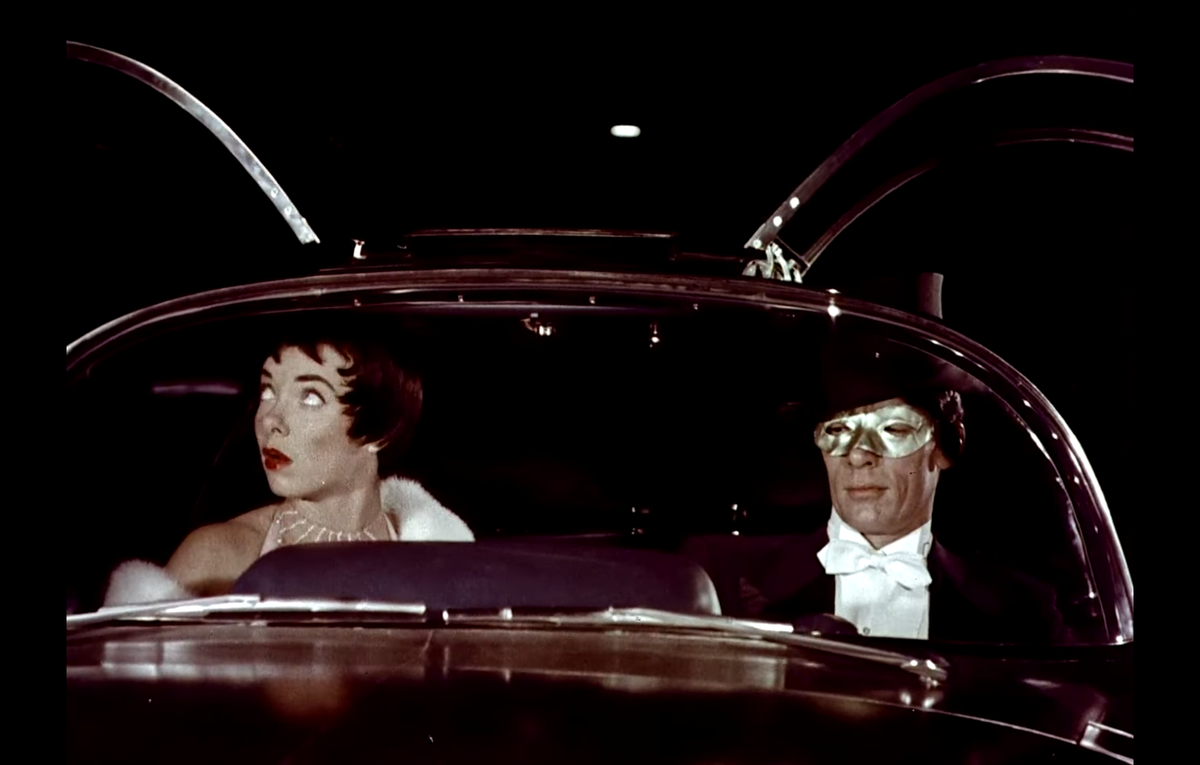 Two people in eveningwear sitting in a futuristic car, with a dark background. 