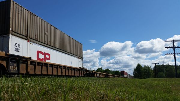 Photo of a train carrying intermodal shipping containers. In the foreground, a grassy field. In the background, a blue sky with fluffy clouds.