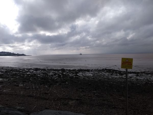 Photo of a beach and a bright but cloudy sky. There is a ship far away on the horizon, and a yellow sign in the foreground that says "Danger Soft Mud."