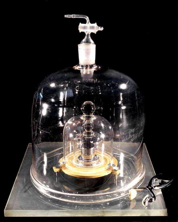 A photo of a small, metal weight within several layers of protective glass cloches. A pair of tongs are placed next to the outermost cloche. The background of the photo is black.