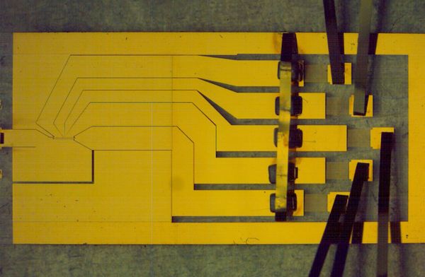 A slightly low-contrast image of a sheet of yellow material with paths cut out of it. At the ends ofthe paths, there are strips of sheet metal sticking up and out.