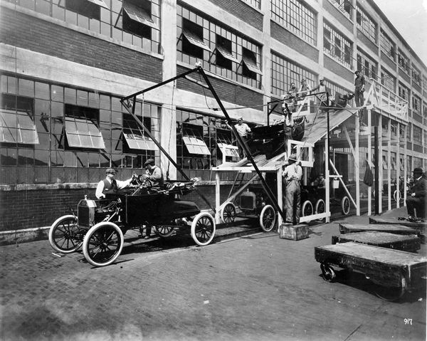 Black and white photo of a large building. On its side, there is a gantry and a ramp. Below the ramp, cars typical of the early-20th century, surrounded by workers.
