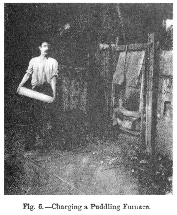 A very grainy, old photo in black and white, depicting a worker carrying a pig of iron to load a furnace.