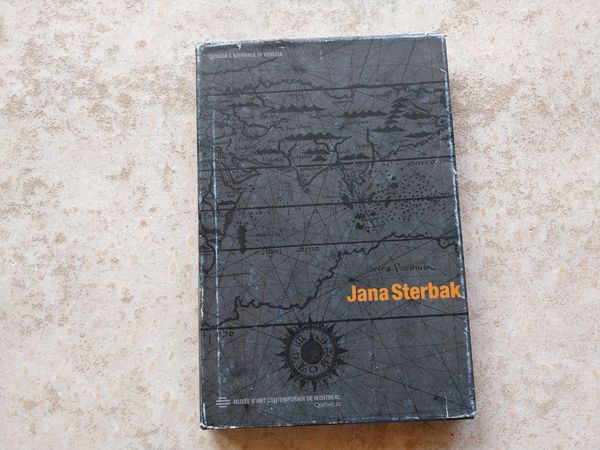 A black book on a concrete background. The book has a map motif, and in orange displays the name "Jana Sterbak."