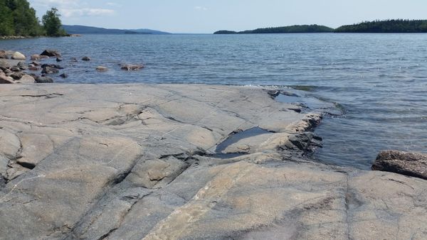 The rocky shore of a lake. The water is a steel-blue shade, and one massive, flat rock is jutting out into the lake.