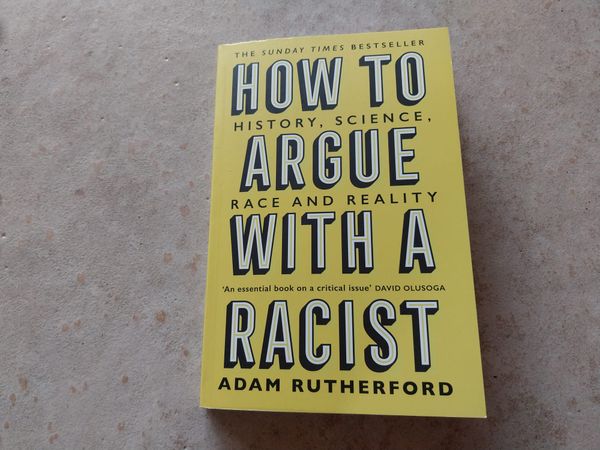 A yellow book on a concrete background. The book is How to Argue with a Racist by Adam Rutherford.