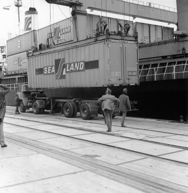 Black-and-white photo of an early shipping container being moved from a ship to a truck, with workers looking on.