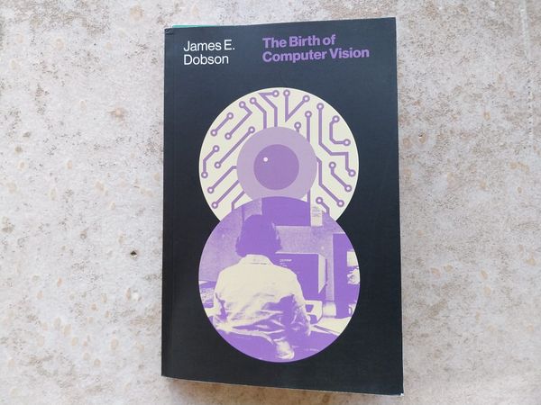 On a concrete floor, a book. The book is The Birth of Computer Vision by James E. Dobson. It has a black cover with some purple-tinted, slightly abstract images representing computation.