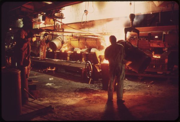 A photo of workers pouring hot metal into moulds. The image is mostly dark, with areas lit up by the heat of the molten metal.