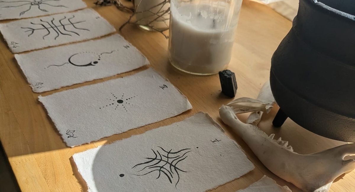 Small cards of parchment paper painted in black ink with sparse sigil designs are laid out on a wood table, to the right of the frame extending back are bones, a cauldron, and candles