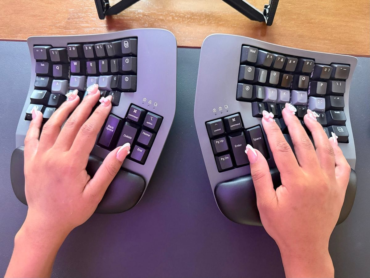 Two hands with pink nails on a split ergonomic keyboard, from above