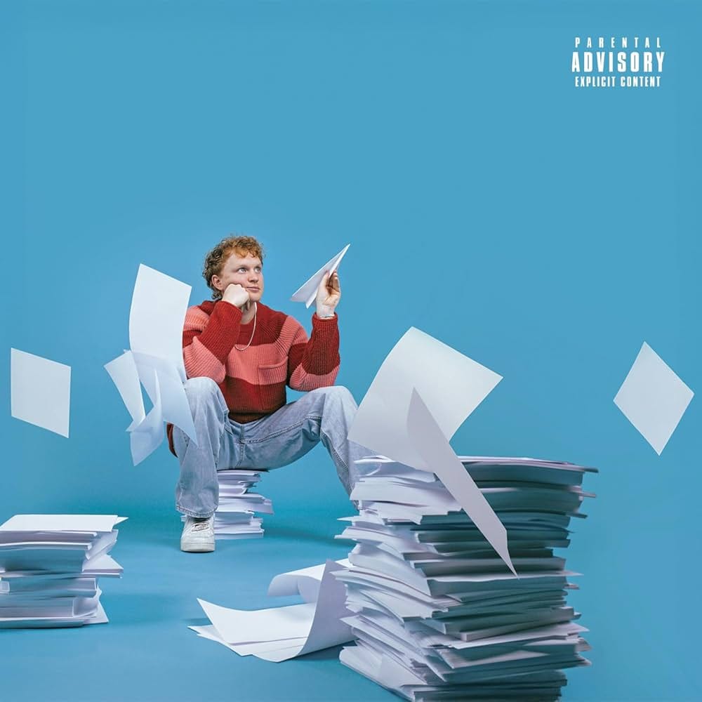 The cover of Knox's "Going, Going, Gone" depicting the singer in a red sweater sitting on a stack of paper, surrounded by other stacks of paper. Some pieces of paper are blowing around. The singer is holding a paper airplane. All of this is in front of a stark blue background.