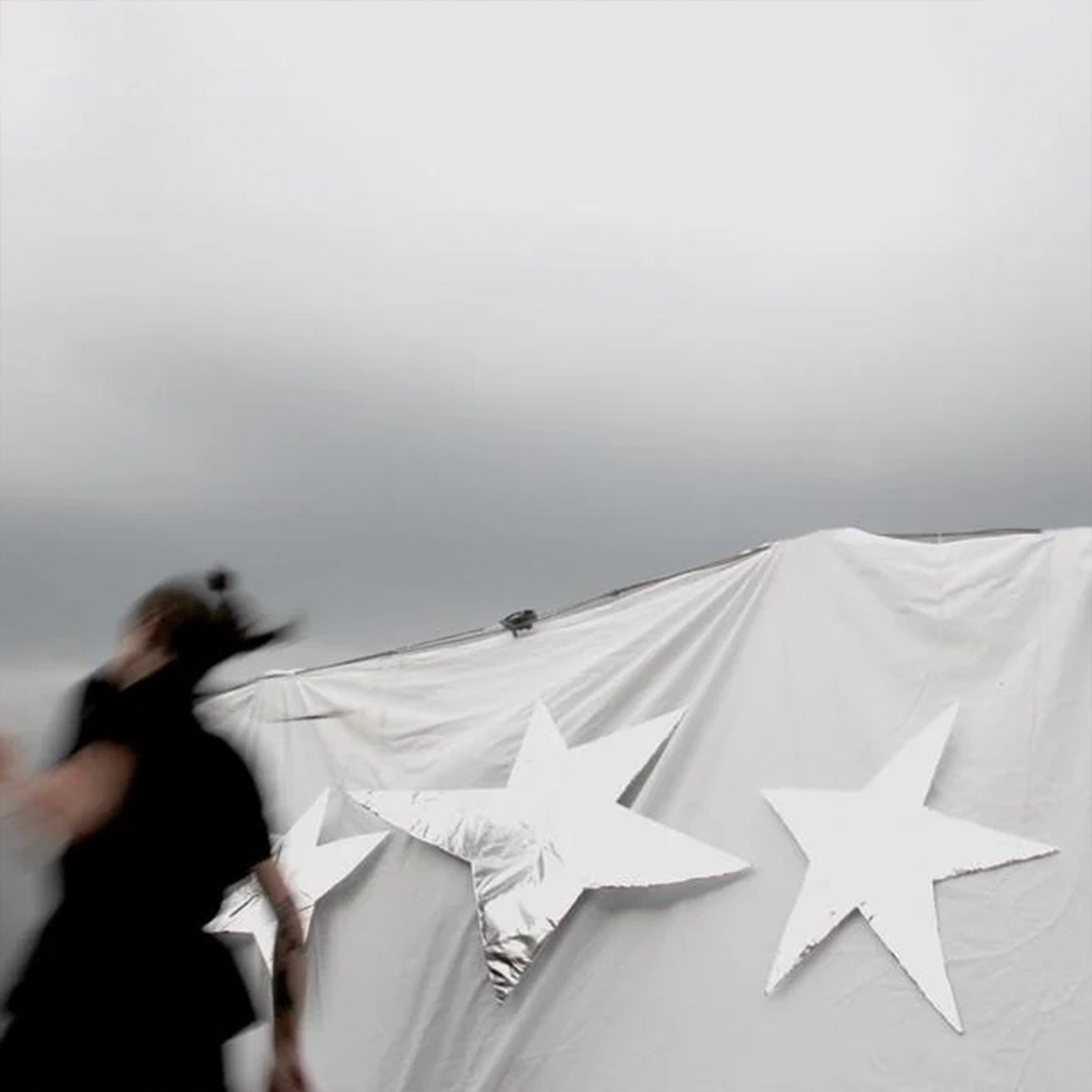 The cover of Young Culture's "All Weapons Formed Against Me Have Prospered" depicting a blurry person in the foreground in front of a sheet with foil stars in front, with a grey sky in the background
