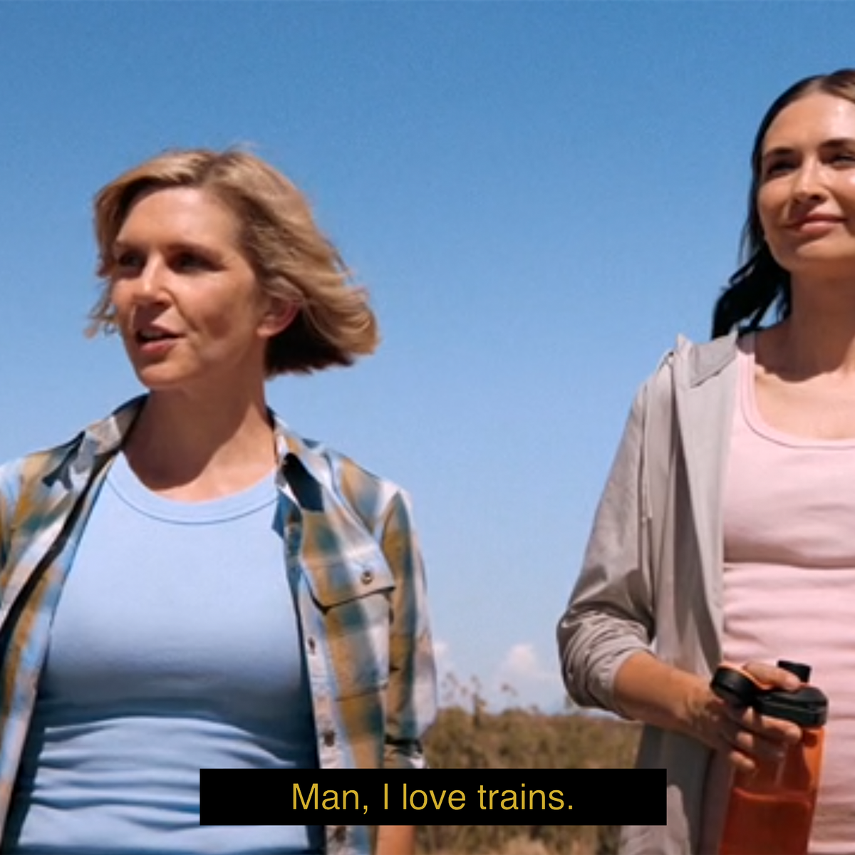 A screenshot of a scene from Pluribus, featuring two women (Carol and Zosia) in the desert. The subtitle reads "Man, I love trains."