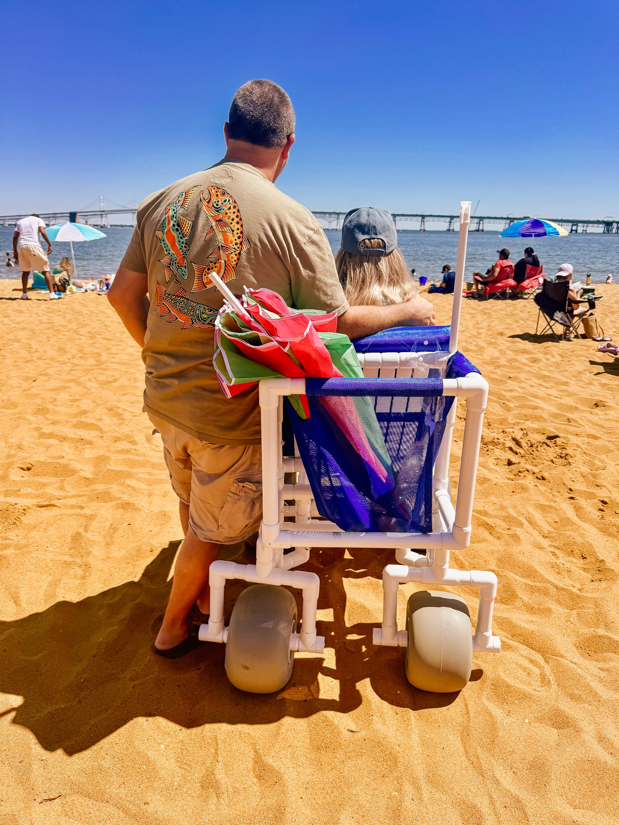 Beach wheelchair user and husband standing next to her, facing the beach on the sand