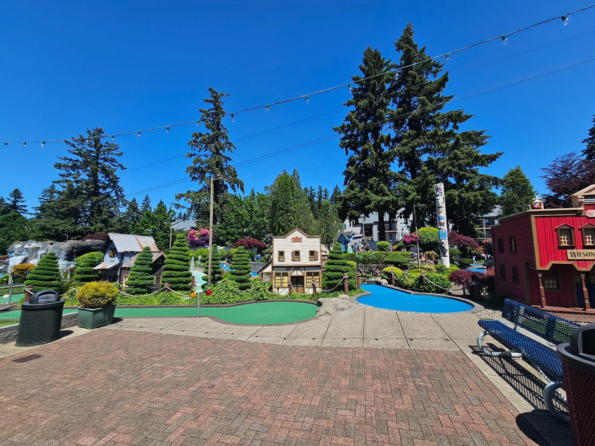 Mini golf course at Bulwinkle's Family Fun Center in Wilsonville, Oregon, with themed buildings, manicured shrubs, a water feature, and string lights on a sunny day
