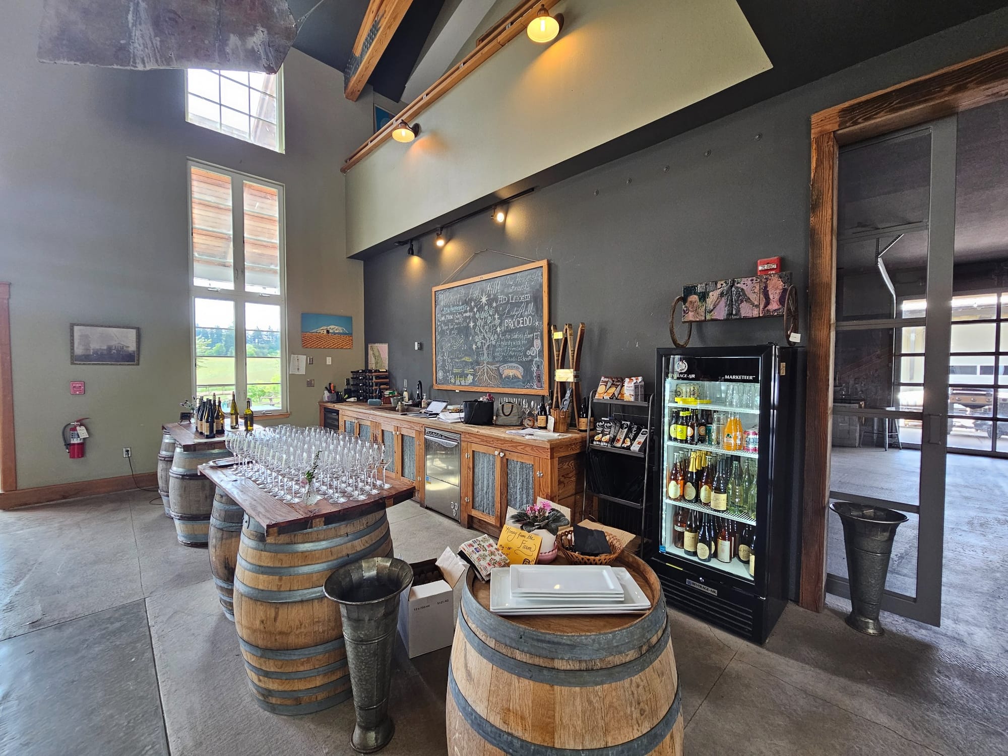 Inside the Lady Hill Winery tasting room in St. Paul, Oregon, with wine barrels used as tasting tables, rows of wine glasses, a chalkboard menu, and high ceilings with natural light