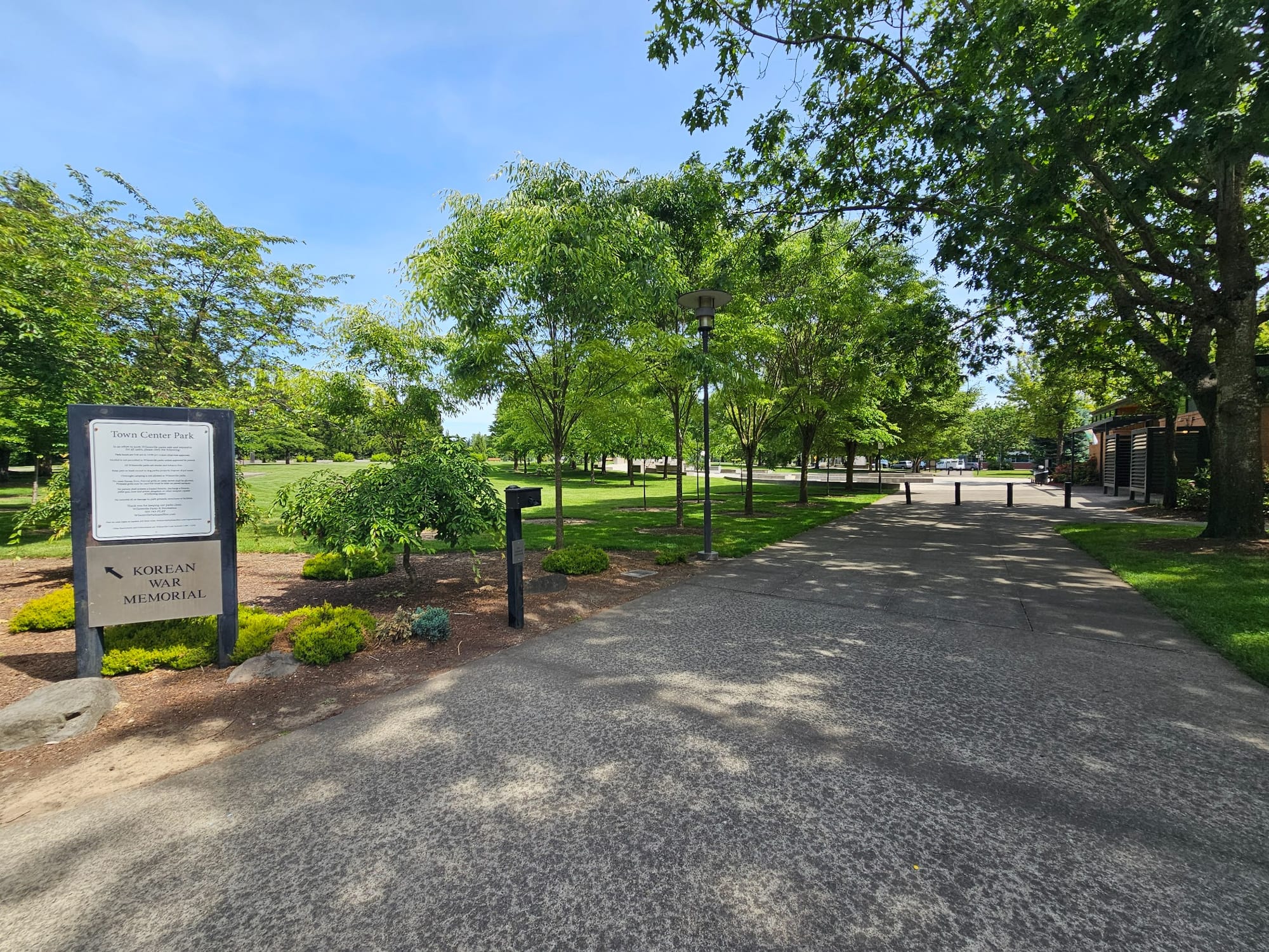 Wide accessible paved path at Town Center Park in Wilsonville, Oregon, with a sign directing visitors to the Korean War Memorial, surrounded by green lawns and trees on a clear summer day